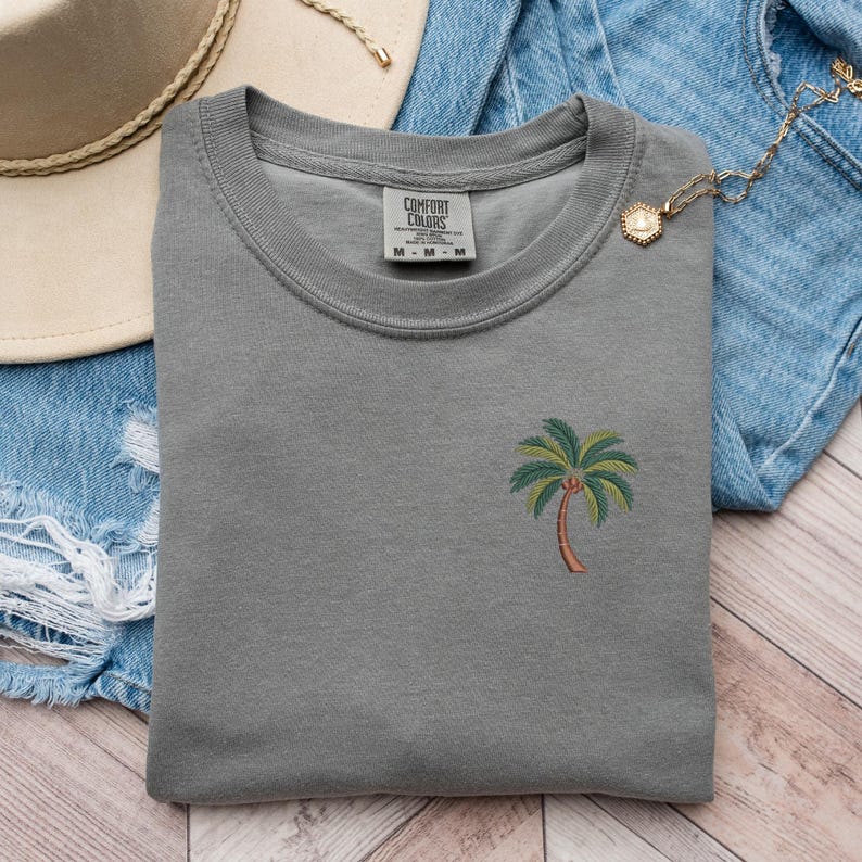 Op de afbeelding: Grijs T-shirt met een klein geborduurd palmboom op de linker borst. Het T-shirt heeft een ronde hals en is gemaakt door Comfort Colors.