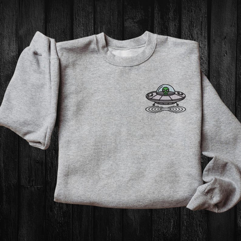 May include: A gray sweatshirt with a small embroidered design of a UFO with an alien inside. The UFO is hovering over a circle of concentric rings.