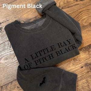 May include: A dark gray sweatshirt with the text "A LITTLE RAY OF PITCH BLACK" in black. The sweatshirt has a small bat graphic on the sleeve. The words "Pigment Black" are at the top of the image.