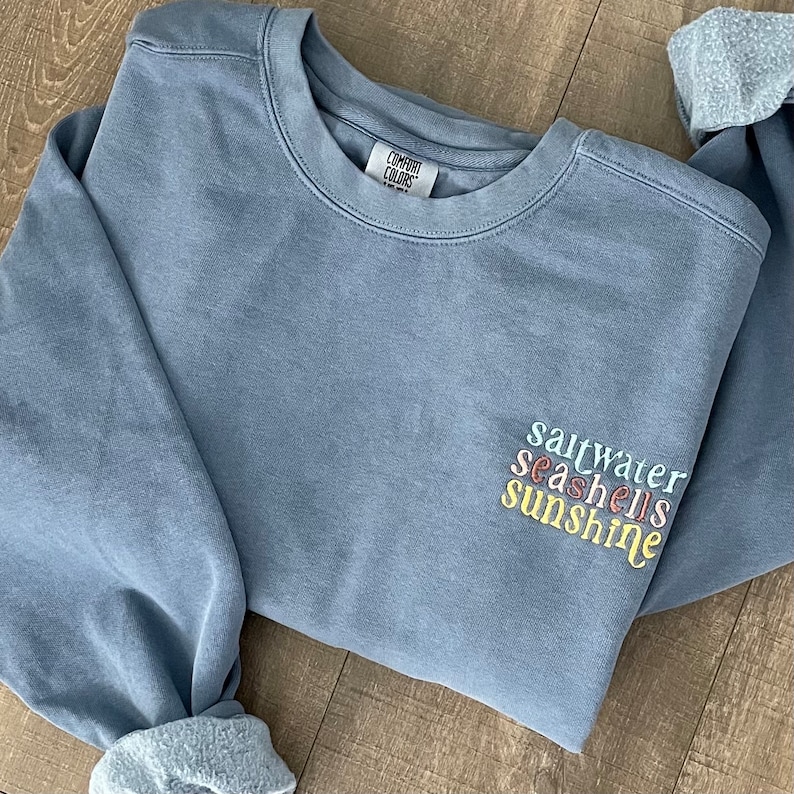 May include: A light blue sweatshirt with the words "saltwater seashells sunshine" embroidered on the front in pastel colors.