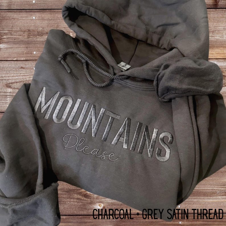 May include: Charcoal gray hoodie with the embroidered text "MOUNTAINS Please" in grey satin thread. The hoodie features a drawstring hood and a front pocket, perfect for casual wear.