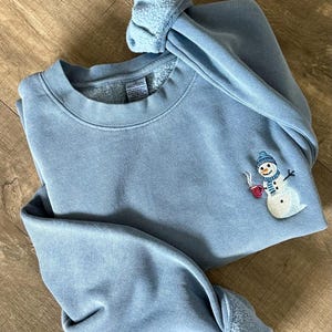 May include: A light blue sweatshirt with a snowman embroidery on the front. The snowman wears a blue hat and scarf and holds a red mug. The cuffs and collar are a lighter shade of blue.