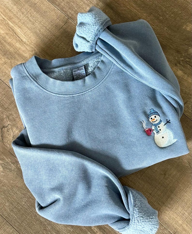 May include: A light blue sweatshirt with a snowman embroidery on the front. The snowman wears a blue hat and scarf and holds a red mug. The cuffs and collar are a lighter shade of blue.