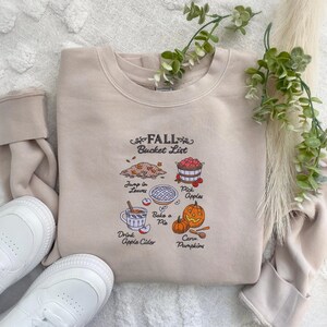 May include: Beige sweatshirt with a "Fall Bucket List" graphic. The graphic includes illustrations of jumping in leaves, picking apples, baking a pie, drinking apple cider, and carving pumpkins. White sneakers are visible.