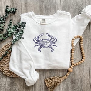 K&ouml;nnte beinhalten: Wei&szlig;es Sweatshirt mit einem marineblauen Krabben-Design. Die Krabbe ist detailliert gestickt, mit deutlich definierten Scheren und K&ouml;rper. Das Sweatshirt liegt auf einer Holzoberfl&auml;che, zusammen mit einer Perlenkette und Dekorationen.