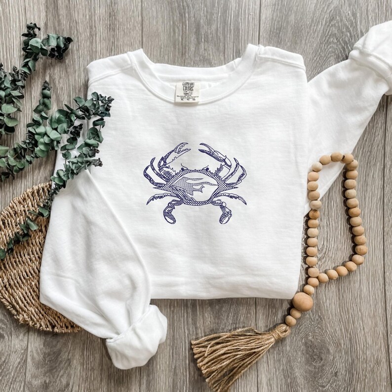 K&ouml;nnte beinhalten: Wei&szlig;es Sweatshirt mit einem marineblauen Krabben-Design. Die Krabbe ist detailliert gestickt, mit deutlich definierten Scheren und K&ouml;rper. Das Sweatshirt liegt auf einer Holzoberfl&auml;che, zusammen mit einer Perlenkette und Dekorationen.