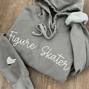May include: Gray hoodie with the words "Figure Skater" in white script. The left sleeve features a small embroidered ice skate. The hoodie has a drawstring hood and a soft, fleecy interior.