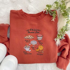 May include: Rust-colored sweatshirt with a Fall Bucket List graphic. The graphic includes illustrations of leaves, apples, pie, pumpkins, and apple cider. The text on the sweatshirt reads "Fall Bucket List".