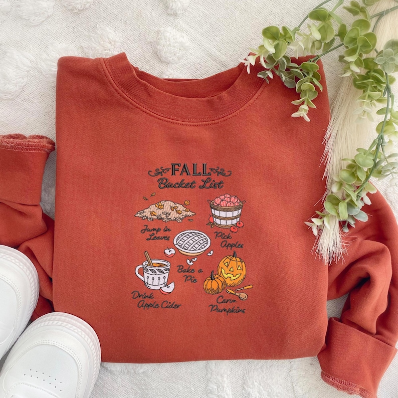May include: Rust-colored sweatshirt with a Fall Bucket List graphic. The graphic includes illustrations of leaves, apples, pie, pumpkins, and apple cider. The text on the sweatshirt reads "Fall Bucket List".