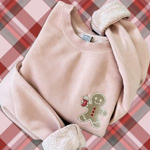 May include: A light pink sweatshirt with a gingerbread man design holding a coffee cup. The gingerbread man has a smiling face and red buttons. The cuffs are folded over, revealing a soft, white lining. The background is a red and pink plaid pattern.