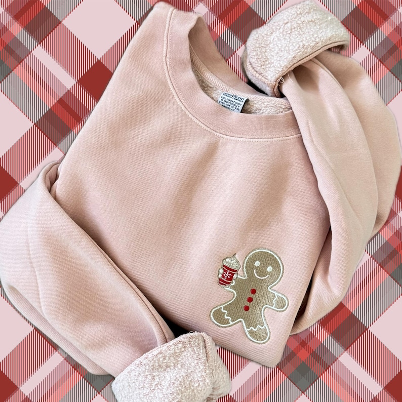 May include: A light pink sweatshirt with a gingerbread man design holding a coffee cup. The gingerbread man has a smiling face and red buttons. The cuffs are folded over, revealing a soft, white lining. The background is a red and pink plaid pattern.