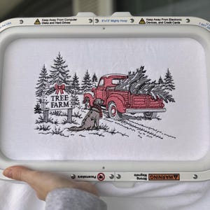 Może przedstawiać: Wzór haftu na białej tkaninie w tamborku 20x33 cm przedstawiający czerwony zabytkowy samochód ciężarowy przewożący sosnę, psa w szaliku oraz znak z napisem TREE FARM i czerwoną kokardą. Scena zawiera czarne drzewa iglaste.