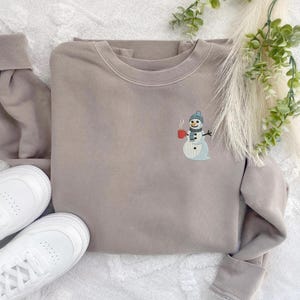 May include: A taupe sweatshirt with a crew neck and long sleeves. A small embroidered snowman wearing a blue hat and scarf, holding a red mug, is on the front. White sneakers are visible on the left side of the image.