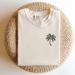 Op de afbeelding: Een cr&egrave;mekleurig T-shirt met een groene palmboom geborduurd op de voorkant. Het T-shirt heeft een ronde hals en de tekst "Cotton Colors" en "S - P - CH" staat gedrukt op het label.