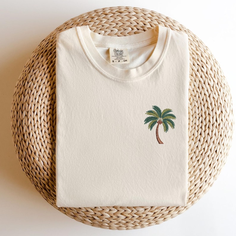 Op de afbeelding: Een cr&egrave;mekleurig T-shirt met een groene palmboom geborduurd op de voorkant. Het T-shirt heeft een ronde hals en de tekst "Cotton Colors" en "S - P - CH" staat gedrukt op het label.