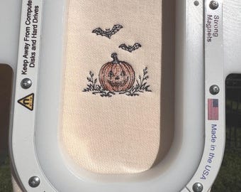 Puede incluir: Una tela de color beige claro asegurada en un bastidor de bordado blanco. La tela presenta un diseño bordado de una calabaza naranja tallada con una cara, rodeada de hierba en trazos negros y dos pequeños murciélagos volando.
