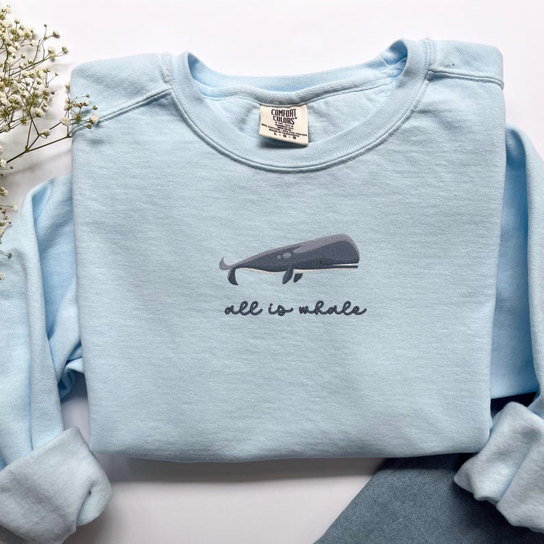 Może przedstawiać: Jasno błękitna bluza z szarym nadrukiem przedstawiającym wieloryba i napisem "all is whale" w kolorze szarym.