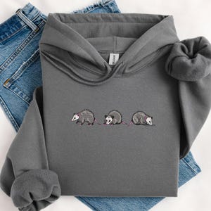 May include: A dark gray hooded sweatshirt with a white embroidered design of three opossums walking in a line.