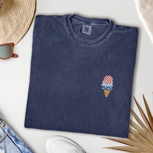 May include: A navy blue t-shirt with a small embroidered ice cream cone design. The ice cream cone features red and white stripes, blue stars, and a tan cone. The shirt is folded and lies on a white surface.