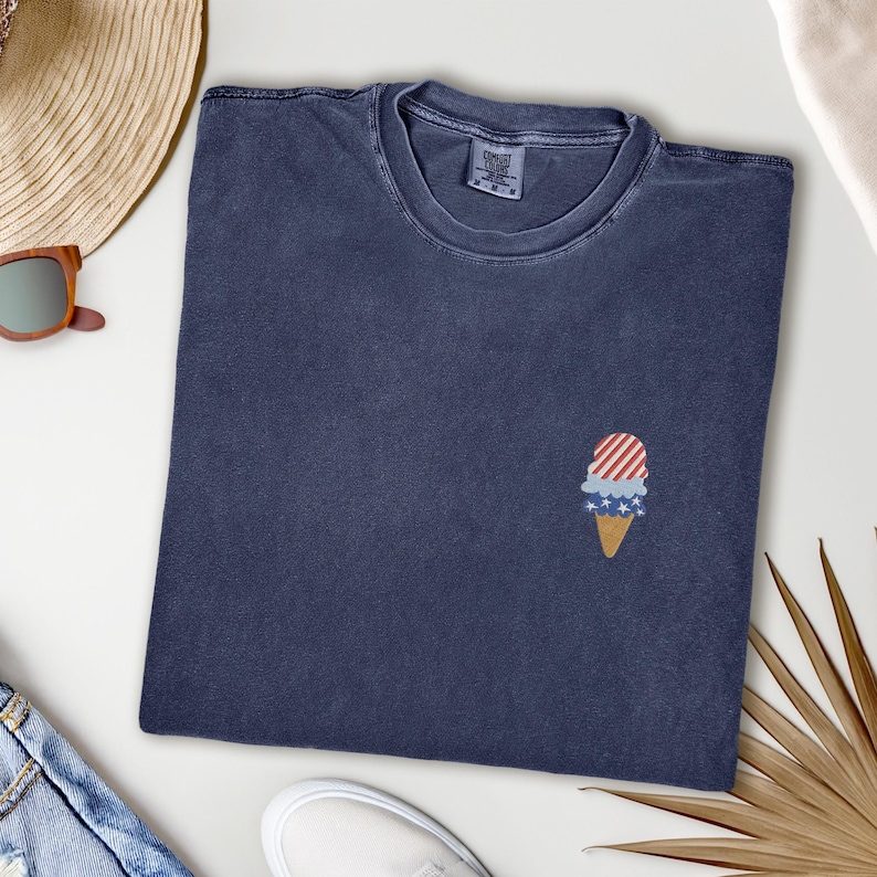 May include: A navy blue t-shirt with a small embroidered ice cream cone design. The ice cream cone features red and white stripes, blue stars, and a tan cone. The shirt is folded and lies on a white surface.