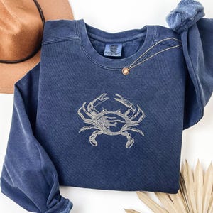 K&ouml;nnte beinhalten: Ein marineblaues Sweatshirt mit einem Krabbenmuster, das in einem helleren Blauton gestickt ist. Das Sweatshirt hat einen Rundhalsausschnitt und lange &Auml;rmel. Eine goldene Halskette mit Anh&auml;nger ist &uuml;ber die Vorderseite drapiert. Ein brauner Hut ist im Hintergrund.