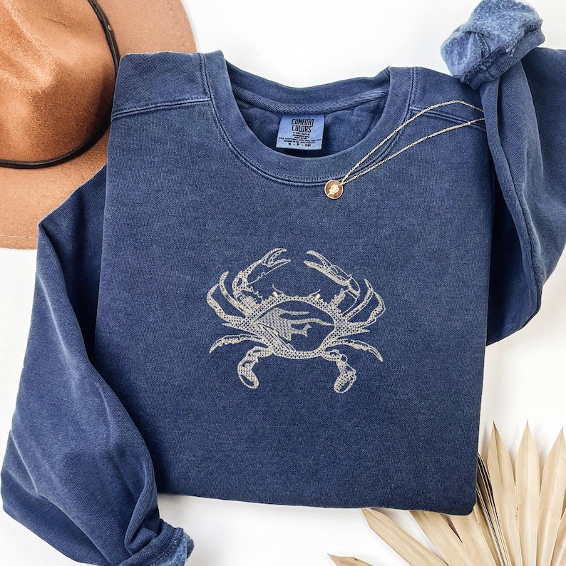 K&ouml;nnte beinhalten: Ein marineblaues Sweatshirt mit einem Krabbenmuster, das in einem helleren Blauton gestickt ist. Das Sweatshirt hat einen Rundhalsausschnitt und lange &Auml;rmel. Eine goldene Halskette mit Anh&auml;nger ist &uuml;ber die Vorderseite drapiert. Ein brauner Hut ist im Hintergrund.