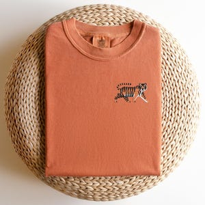 May include: A rust-colored t-shirt with an embroidered tiger design. The t-shirt is folded and displayed on a woven straw mat.
