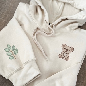 May include: A cream-colored hoodie with a green leaf design on one sleeve and a brown koala design on the other sleeve.