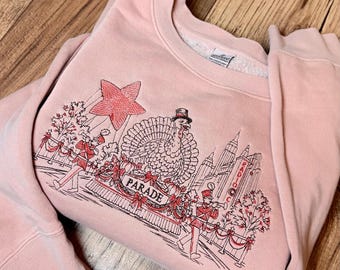 Peut inclure: Un sweat-shirt à col rond rose poudré avec une broderie représentant une parade de Thanksgiving. Le motif inclut une dinde portant un haut-de-forme, des musiciens de fanfare, une grande étoile et un panneau avec le texte PARADE et RADIO.