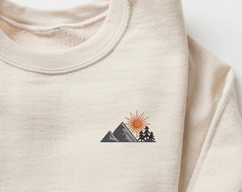 Puede incluir: Una sudadera de color crema con un pequeño diseño bordado de una cordillera, un sol y árboles.