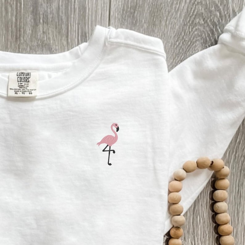 May include: White long-sleeve shirt with a small pink flamingo embroidered on the chest. The flamingo has a black beak and legs. The shirt has a tag that says "Comfort Colors".