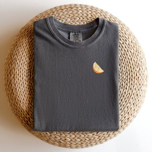 Puede incluir: Una camiseta gris oscuro con una peque&ntilde;a rodaja de naranja bordada en la parte delantera.