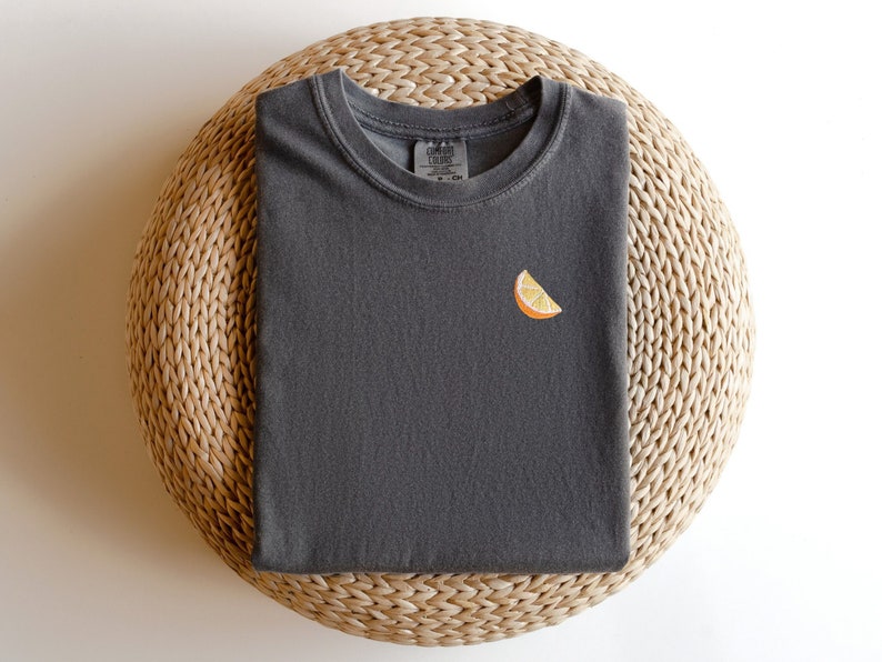 Puede incluir: Una camiseta gris oscuro con una peque&ntilde;a rodaja de naranja bordada en la parte delantera.