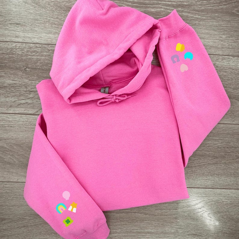 Peut inclure: Un sweat &agrave; capuche rose avec des patchs brod&eacute;s color&eacute;s sur les manches. Les patchs comprennent un arc-en-ciel, un tr&egrave;fle &agrave; quatre feuilles, un c&oelig;ur, une &eacute;toile et un nuage.