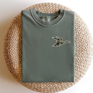 May include: A green t-shirt with a camouflage duck design on the front. The shirt is folded and sitting on a woven basket.