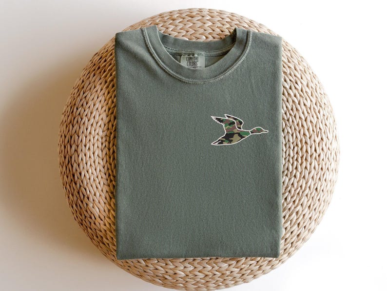 May include: A green t-shirt with a camouflage duck design on the front. The shirt is folded and sitting on a woven basket.