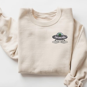 May include: A beige sweatshirt with a small embroidered image of a UFO with a green alien inside. The UFO is hovering over a circle of ripples.