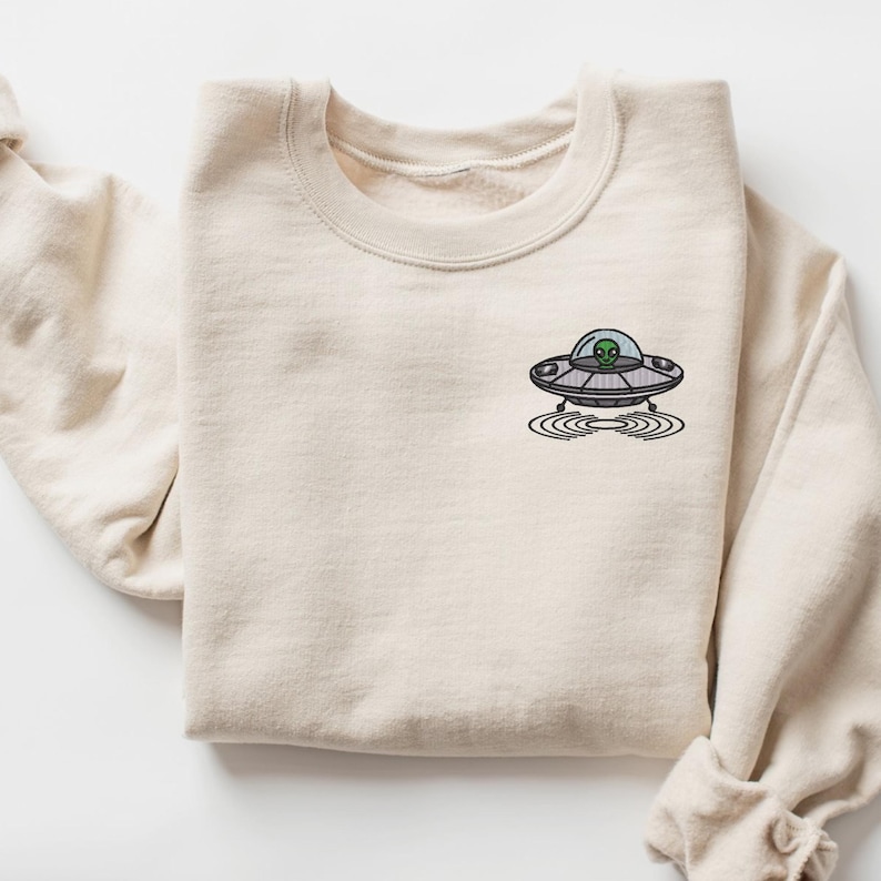 May include: A beige sweatshirt with a small embroidered image of a UFO with a green alien inside. The UFO is hovering over a circle of ripples.
