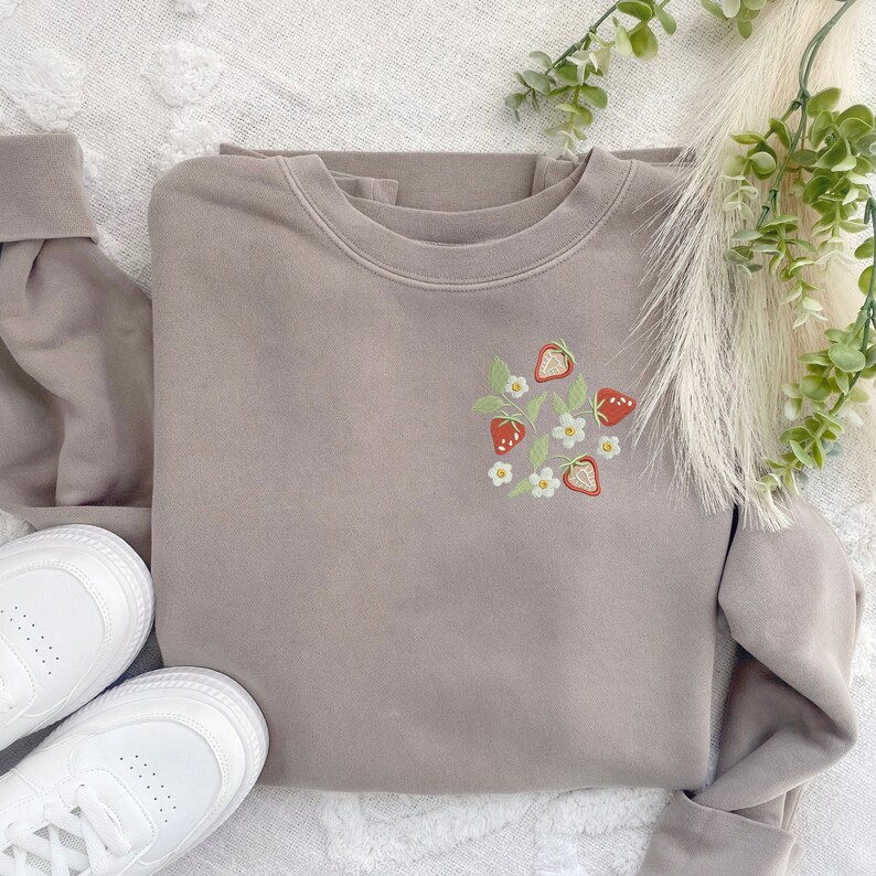 May include: A light brown sweatshirt with an embroidered design of strawberries and white flowers on the front.