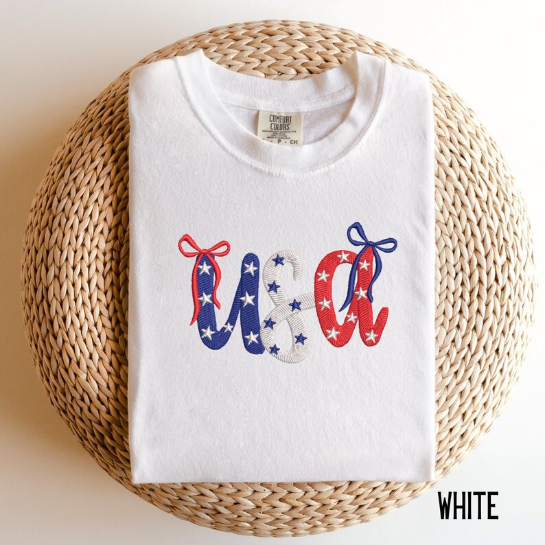 Pu&ograve; includere: T-shirt bianca con "USA" ricamato in rosso, bianco e blu. Le lettere sono decorate con stelle e fiocchi. La maglietta &egrave; piegata su una superficie circolare intrecciata. La parola "WHITE" &egrave; nell'angolo in basso a destra.