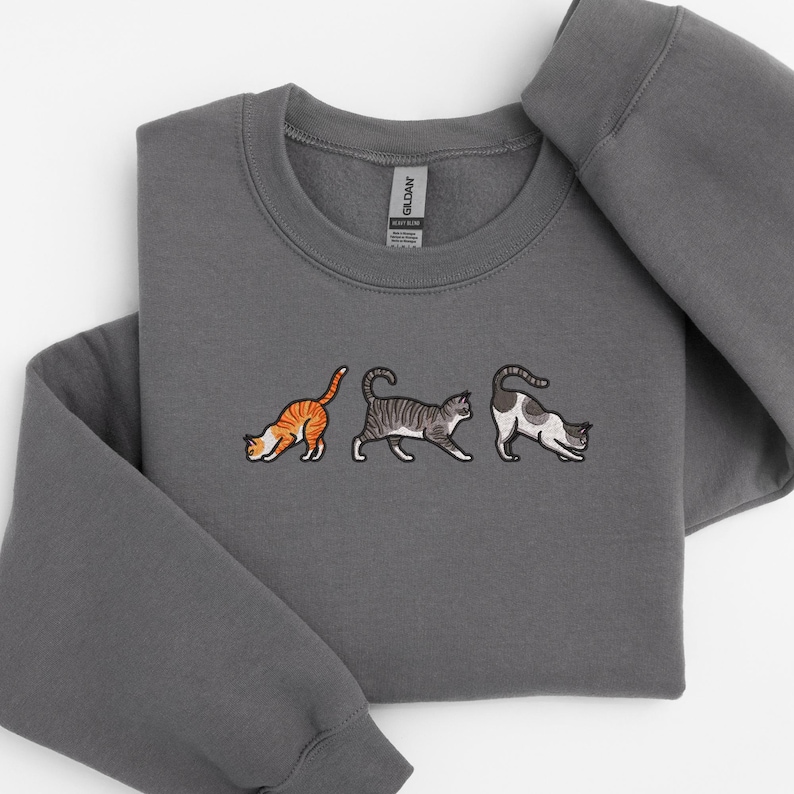 May include: A gray sweatshirt with an embroidered design of three cats walking in a line. The cats are orange, gray and white.