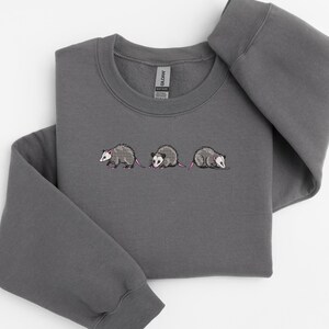 May include: A dark gray sweatshirt with an embroidered design of three opossums walking in a line. The opossums are gray with pink ears and noses.