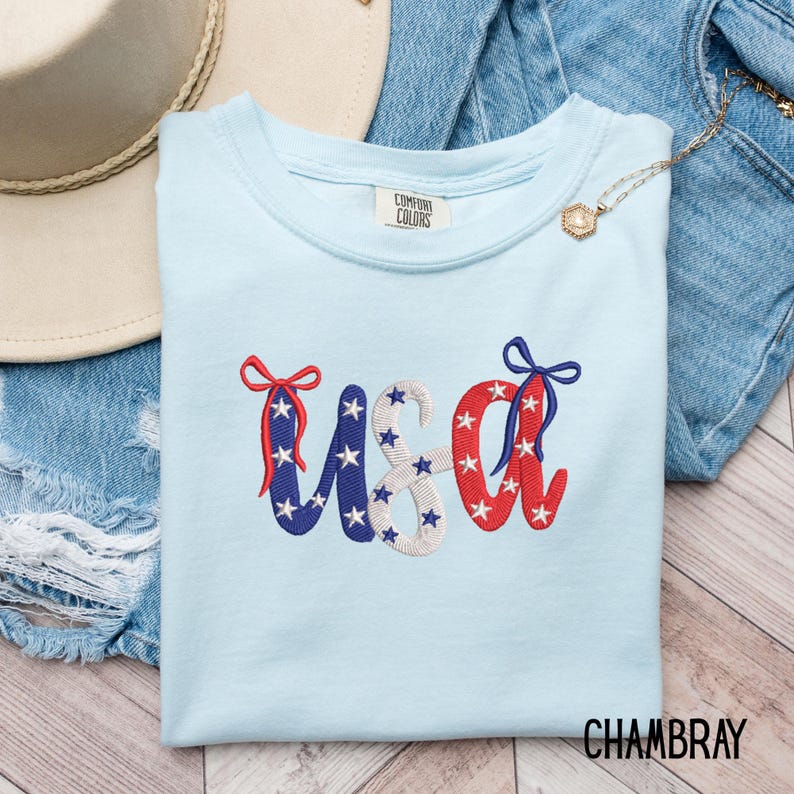 Pu&ograve; includere: Una maglietta azzurra con la scritta "USA" ricamata in rosso, bianco e blu, decorata con stelle e fiocchi. La maglietta &egrave; di Comfort Colors ed &egrave; esposta con pantaloncini di jeans e un cappello di paglia.