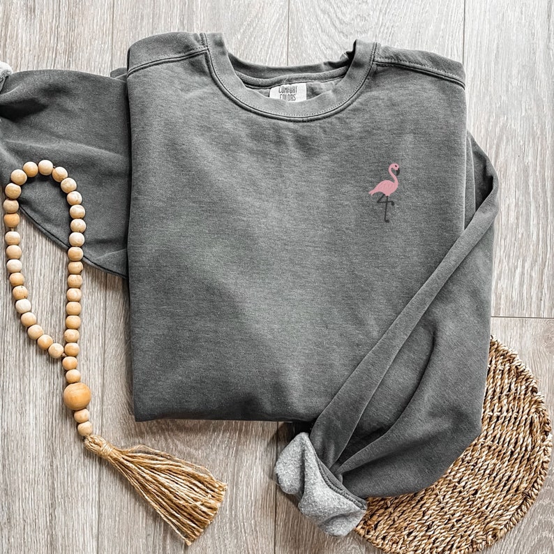 May include: A dark gray sweatshirt with a pink flamingo embroidered on the chest. The sweatshirt has a crew neck and long sleeves. The brand name "Comfort Colors" is printed on the tag. A wooden bead garland and a woven placemat are also in the image.