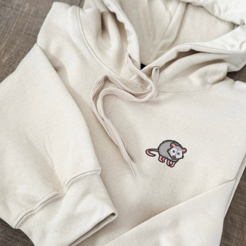 May include: A beige hoodie with a small embroidered possum on the front. The hoodie has a drawstring hood and a kangaroo pocket.