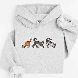 May include: A gray hoodie with a graphic of three cats in different poses. The cats are in a line, with the first cat looking down, the second cat looking forward, and the third cat looking to the right.