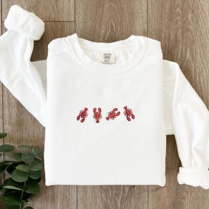 May include: White long-sleeve sweatshirt with four embroidered red lobsters across the chest. The sweatshirt has a crew neck and long sleeves. The brand tag is visible on the upper right side of the shirt.