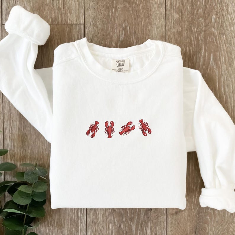 May include: White long-sleeve sweatshirt with four embroidered red lobsters across the chest. The sweatshirt has a crew neck and long sleeves. The brand tag is visible on the upper right side of the shirt.