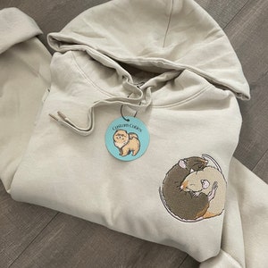 May include: A beige hoodie with a small blue circle sticker that says "Custom Cuddles" and a brown and white embroidered design of two rats cuddling.