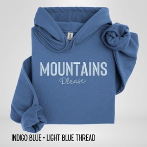 May include: A blue hooded sweatshirt with the words "MOUNTAINS Please" printed on the front in white. The sweatshirt is folded, with one sleeve rolled up. The text "INDIGO BLUE + LIGHT BLUE THREAD" is at the bottom.